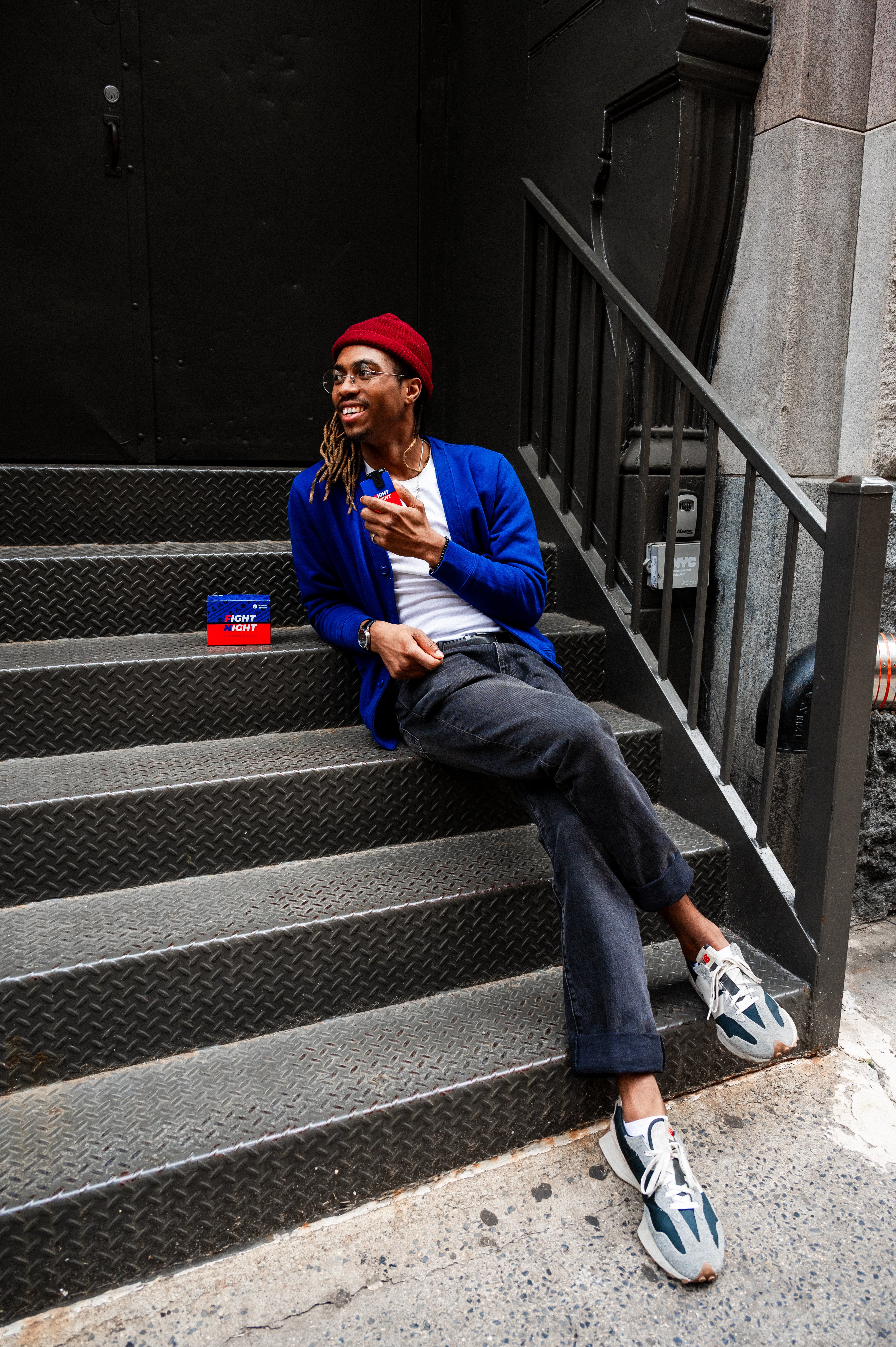 Man sitting on stairs wearing a blue cardigan and red beanie, holding a Fight Night game card in his hand.